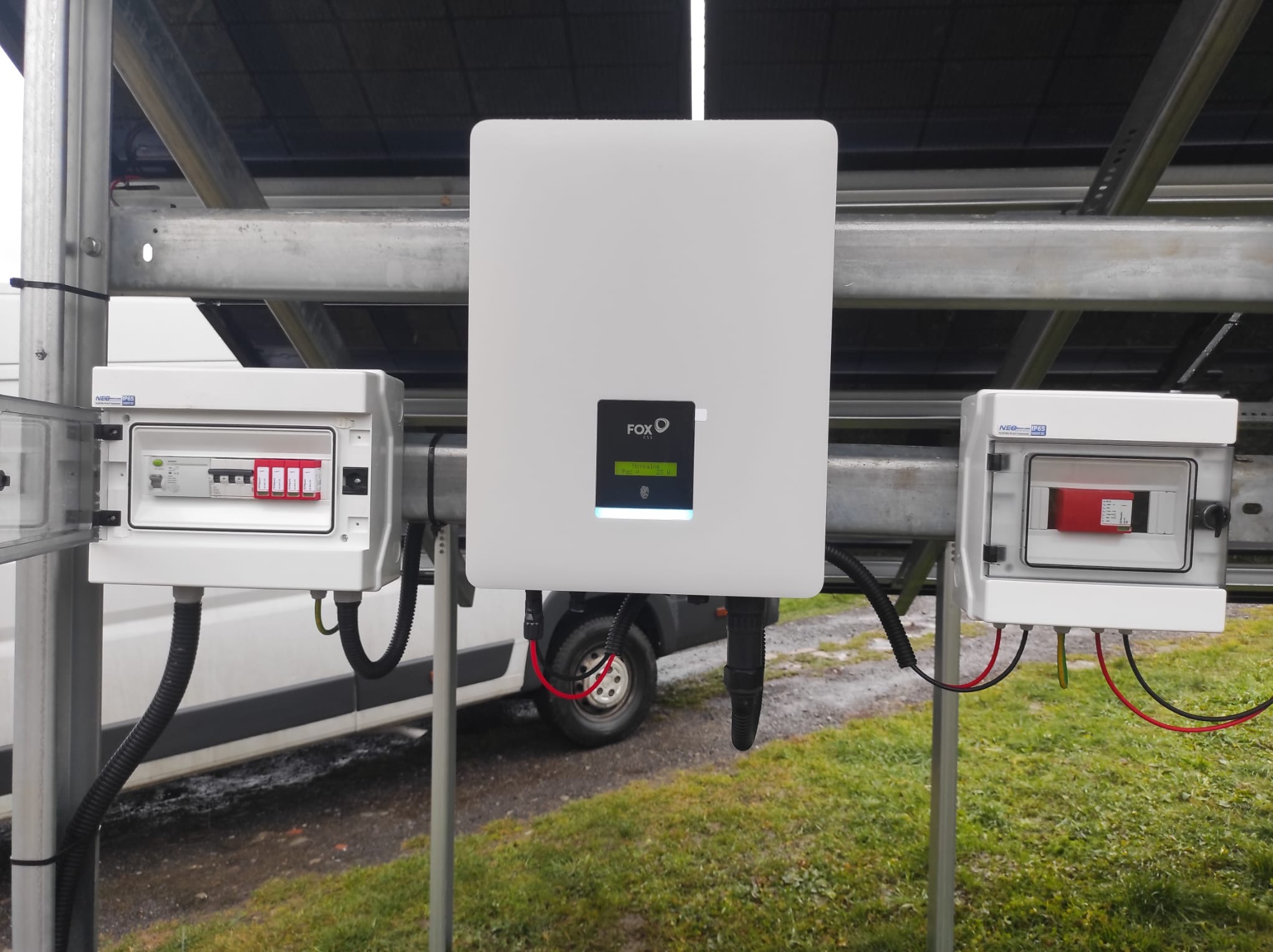 Biały falownik Fox ESS zamontowany na metalowej konstrukcji, z widocznymi skrzynkami elektrycznymi po bokach i instalacją fotowoltaiczną w tle, na tle zielonej trawy i samochodu dostawczego.
