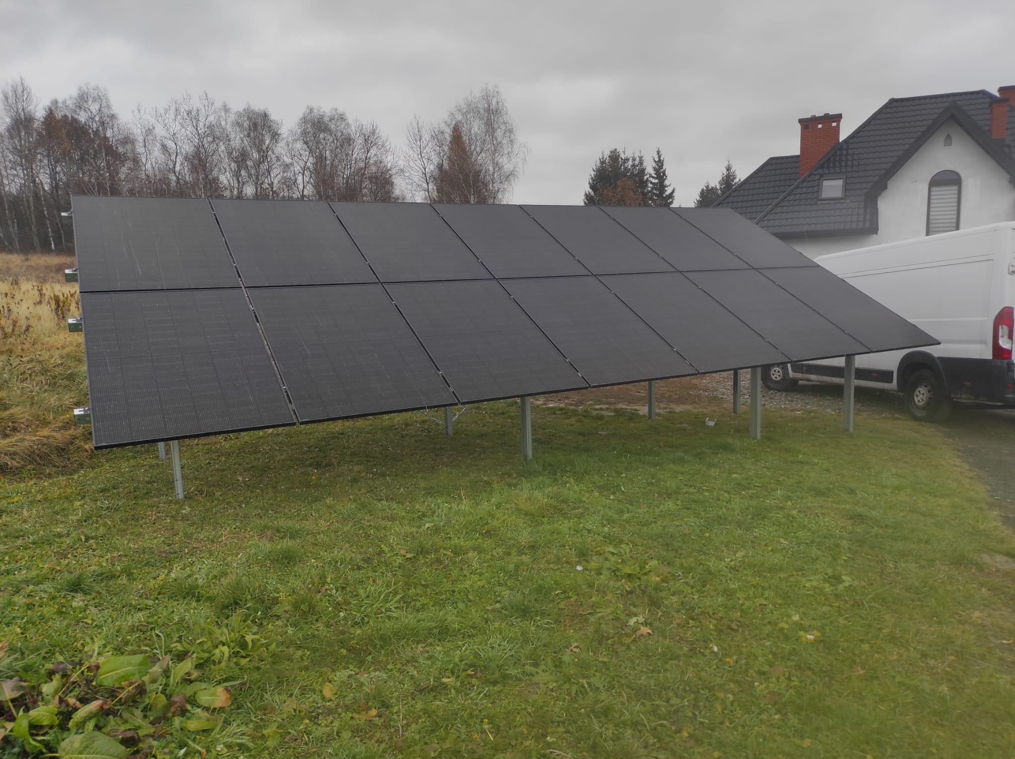 Czarny panel słoneczny na metalowych wspornikach, zainstalowany na trawniku przed domem z białymi ścianami i ciemnym dachem, obok zaparkowany biały bus.