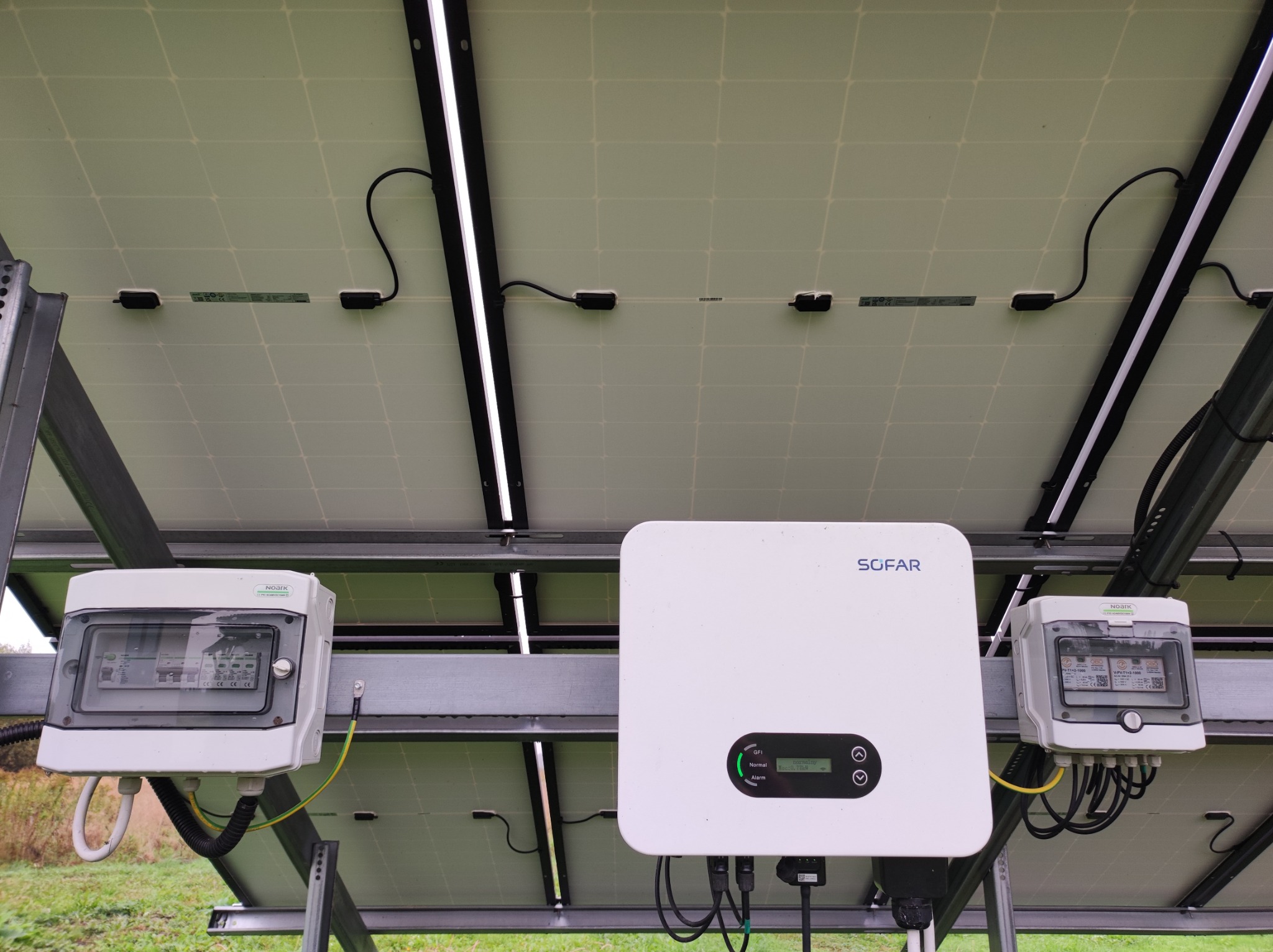 Widok od dołu na panele słoneczne z widocznym inwerterem Sofar i skrzynkami zabezpieczeń NOARK, zamontowane na metalowej konstrukcji na tle zieleni.