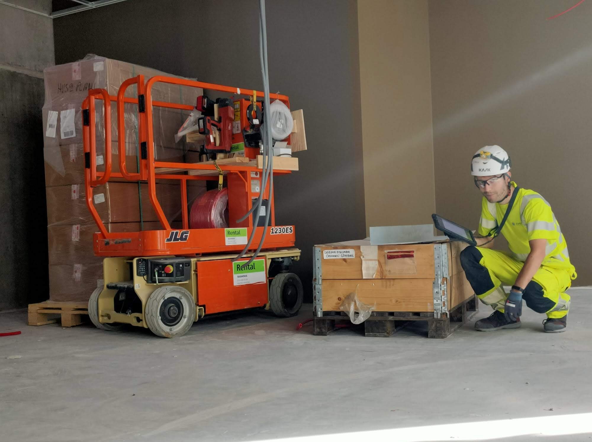 Elektryk w ubraniu roboczym, kuca obok drewnianej skrzyni na palecie, trzymając tablet, obok podnośnik JLG z narzędziami i zwiniętym kablem, w tle kartony na palecie, surowe wnętrze budynku.