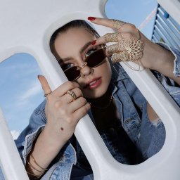 Si Beauty Studio Izabela Sidorska - Portret kobiety w okularach przeciwsłonecznych, z czerwonymi paznokciami, bogatą biżuterią na dłoniach i ustami pomalowanymi błyszczykiem, w denimowej kurtce, pozującej w białej konstrukcji...