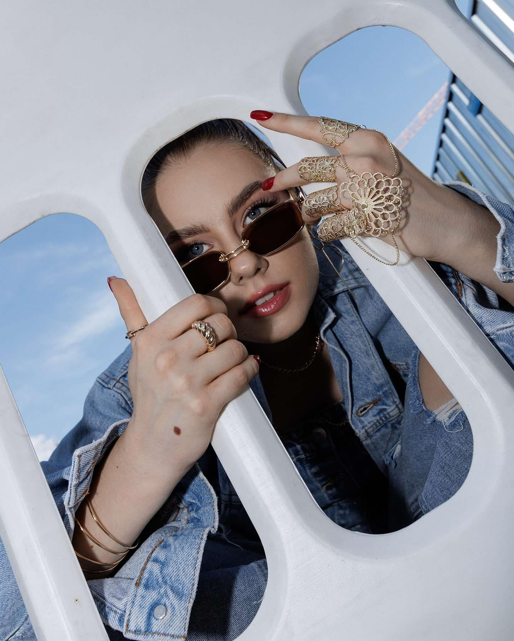 Portret kobiety w okularach przeciwsłonecznych, z czerwonymi paznokciami, bogatą biżuterią na dłoniach i ustami pomalowanymi błyszczykiem, w denimowej kurtce, pozującej w białej konstrukcji...