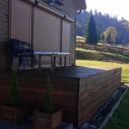 Drewniany dom z tarasem i roletami zewnętrznymi, na tarasie ustawione krzesła i stół, w tle zalesione wzgórza.