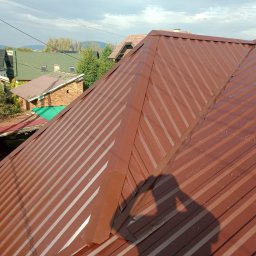 Grzegorz ziolkowski - Nowe, brązowe, blaszane pokrycie dachowe z widocznym cieniem osoby robiącej zdjęcie, w tle zabudowa jednorodzinna i zieleń.