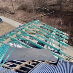 Konstrukcja nowego dachu z pomalowanymi na turkusowo elementami drewnianymi, częściowo pokryta membraną dachową Crysys-X oraz blachą trapezową, widok z góry na tle drzew bez liści.