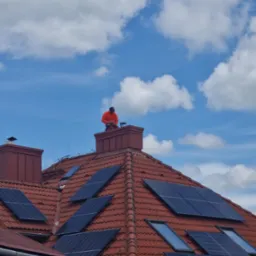 Czerwony dach z panelami słonecznymi i oknami połaciowymi w Milówce. Na kominie dekarz w pomarańczowej kurtce. Błękitne niebo z białymi chmurami w tle.