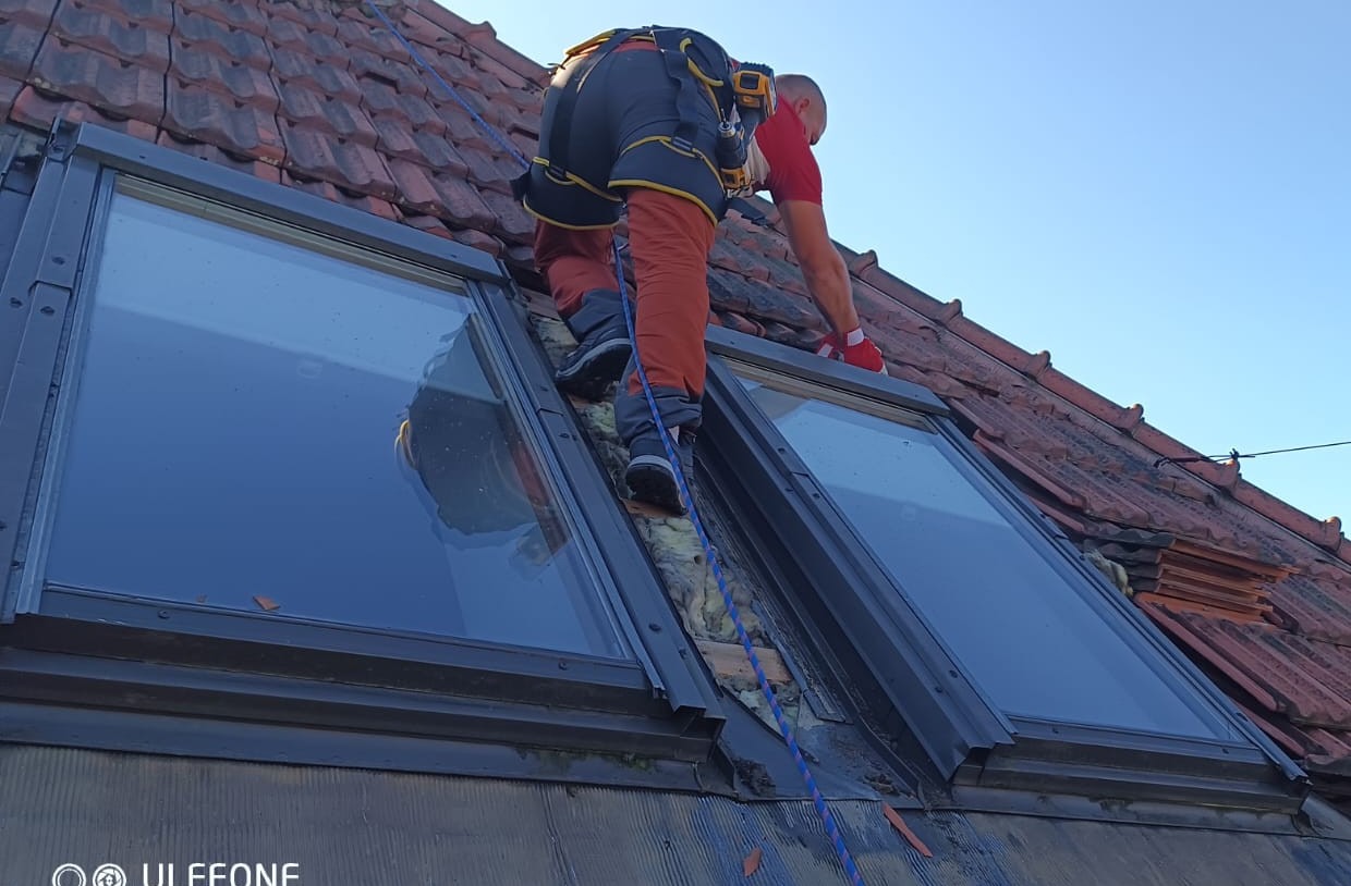 Monter w uprzęży zabezpieczającej, pochylony nad oknem dachowym podczas prac na dachu pokrytym czerwoną dachówką.