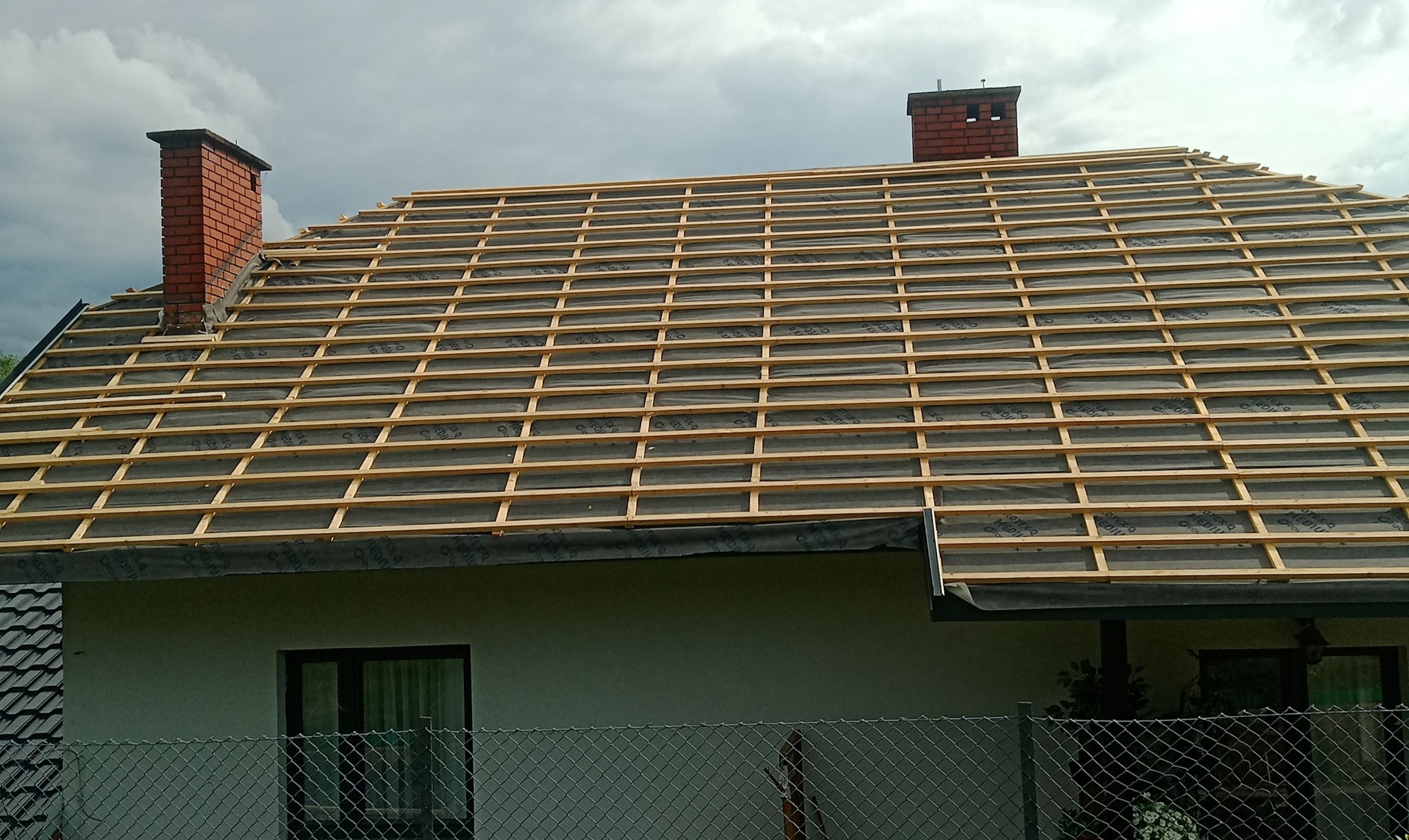 Konstrukcja drewniana łat dachowych ułożona na czarnej membranie dachowej, widoczne kominy z cegły na tle pochmurnego nieba.