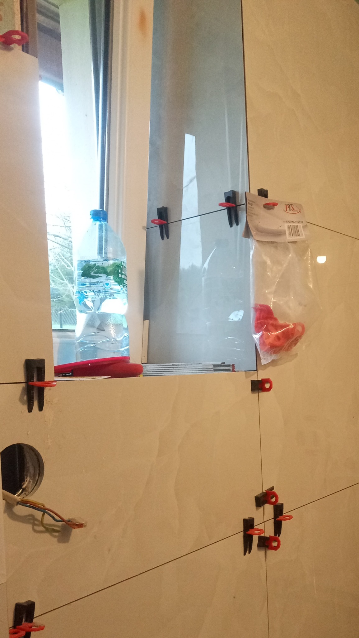 Układanie płytek ceramicznych na ścianie z użyciem systemu poziomowania; widoczna butelka z wodą na parapecie, paczka z klinami i otwór na instalację elektryczną.