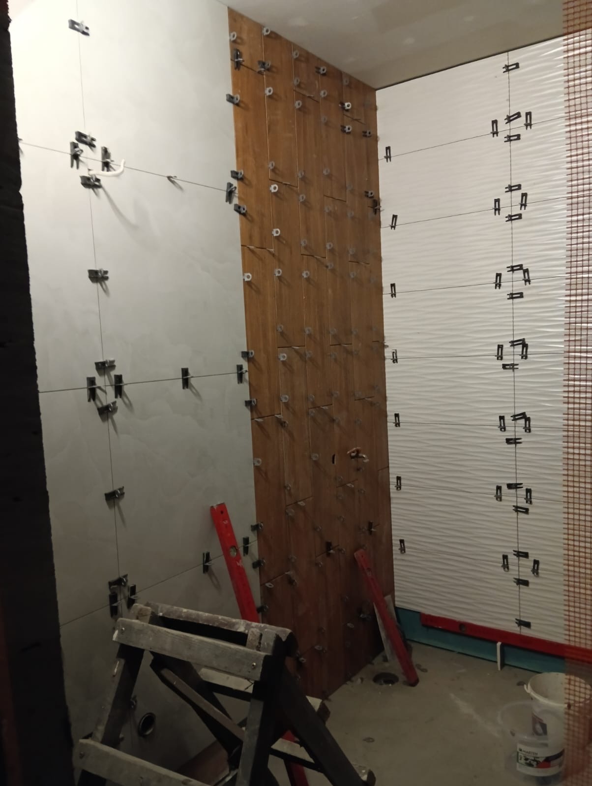 Montaż płytek ściennych w łazience, widoczne różne wzory i kolory płytek, poziomice oraz system poziomowania płytek.