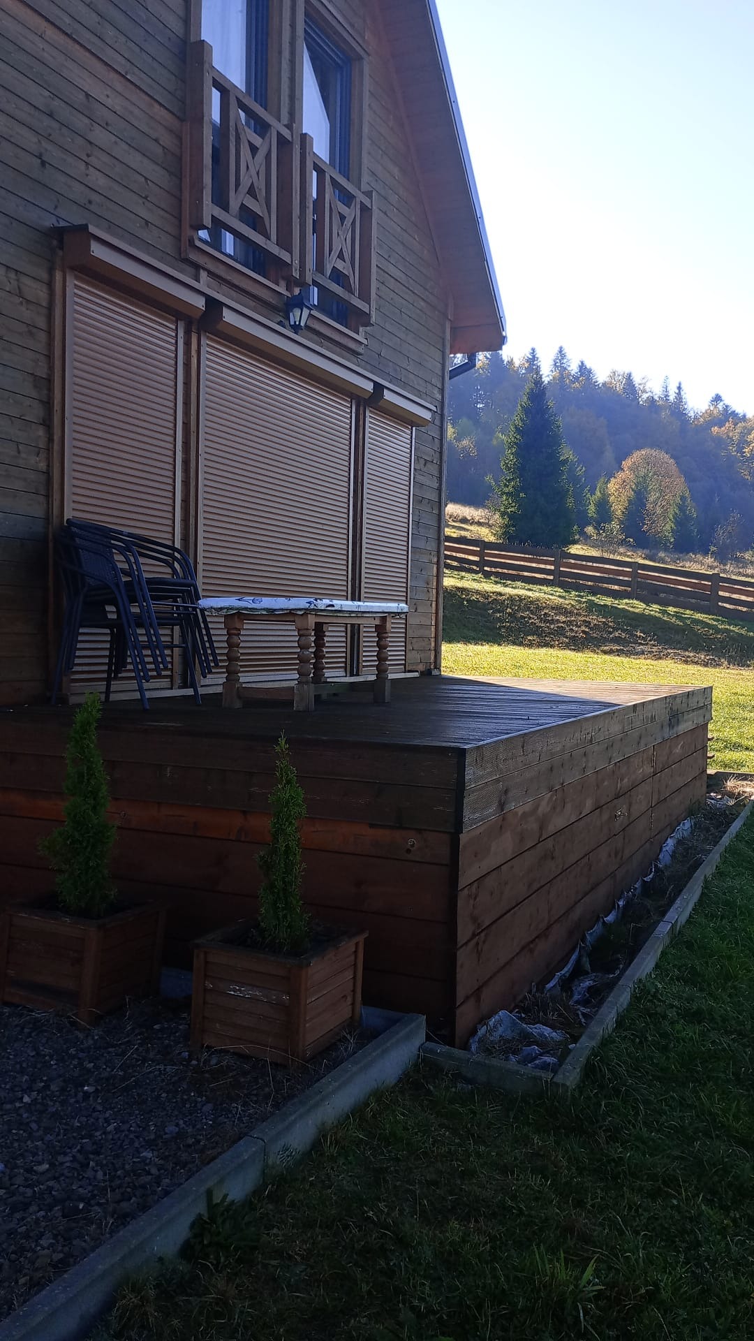 Drewniany dom z tarasem i roletami zewnętrznymi, na tarasie ustawione krzesła i stół, w tle zalesione wzgórza.