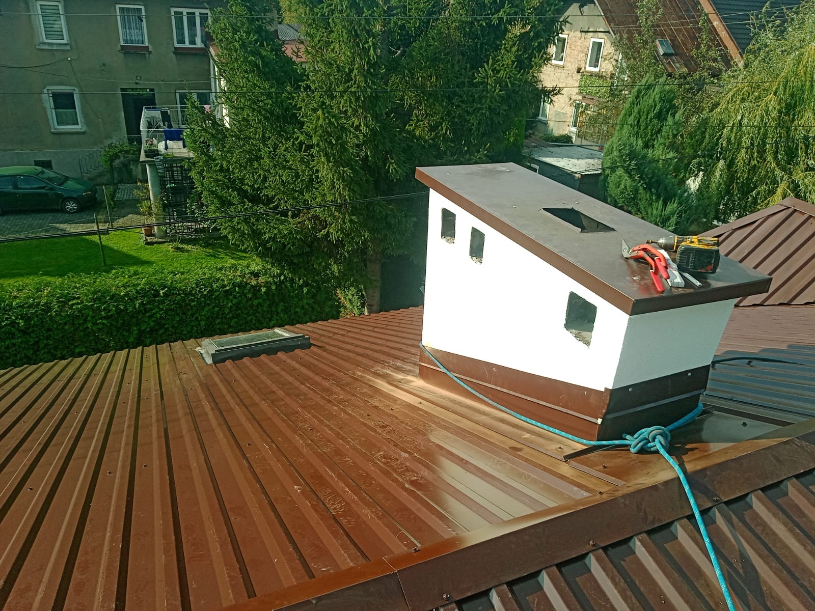 Brązowy blaszany dach z kominem w trakcie obróbki blacharskiej, widoczne narzędzia: wiertarko-wkrętarka i szczypce, zabezpieczony liną, w tle budynki mieszkalne i zieleń.