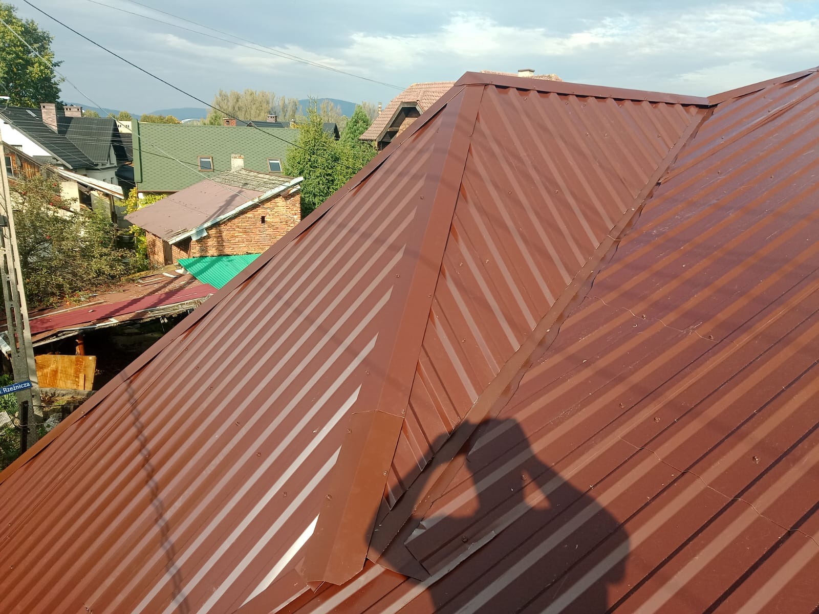 Nowe, brązowe, blaszane pokrycie dachowe z widocznym cieniem osoby robiącej zdjęcie, w tle zabudowa jednorodzinna i zieleń.