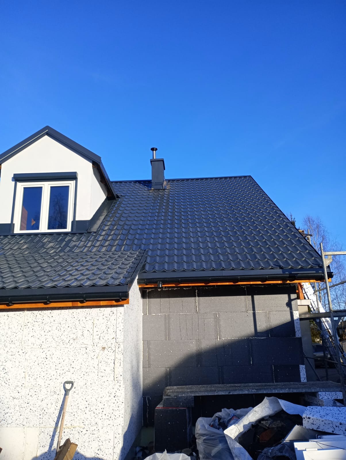 Nowy, ciemnoszary dach z blachodachówki modułowej na domu w trakcie budowy, widoczne orynnowanie, komin i fragment rusztowania.