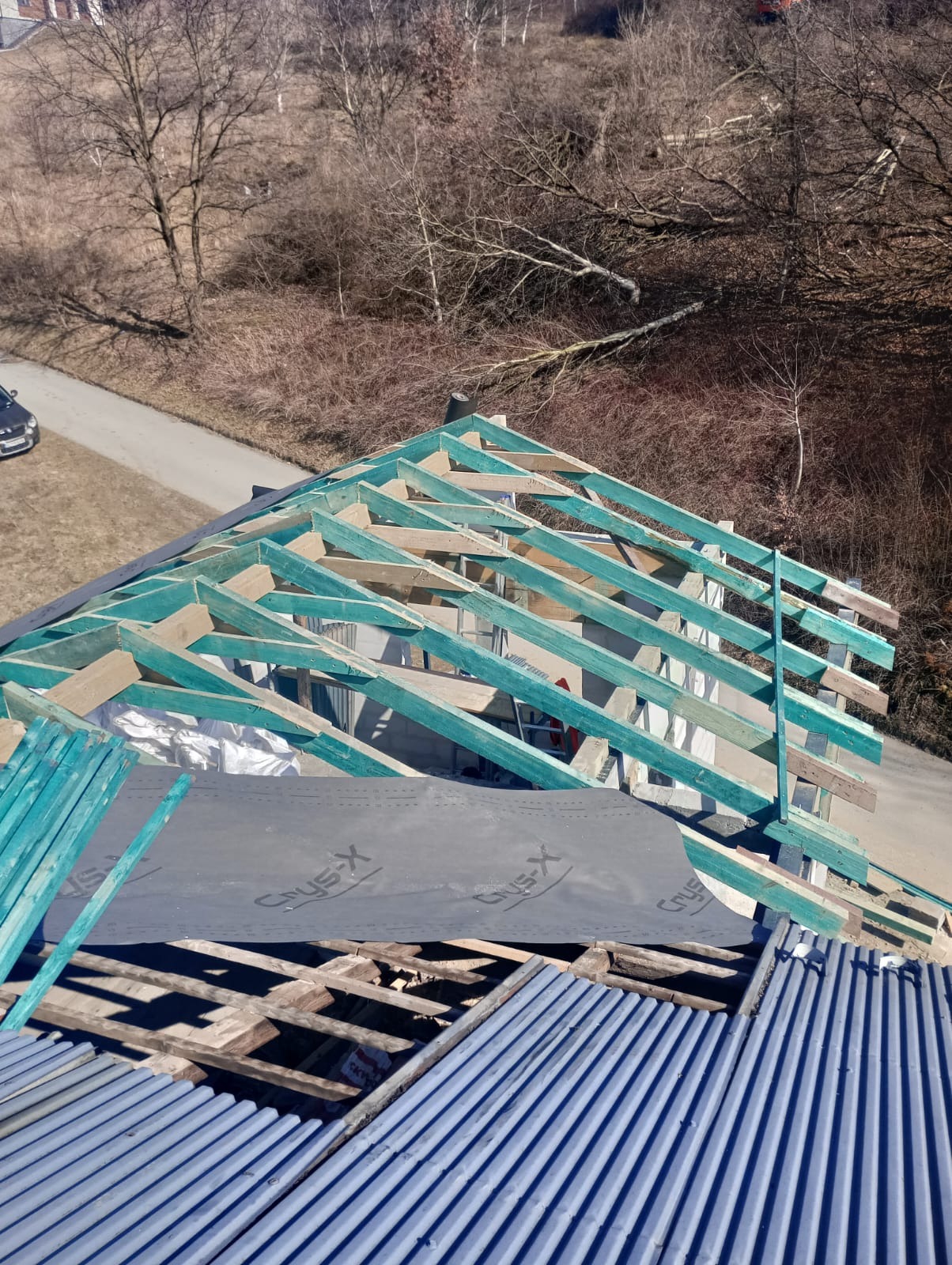 Konstrukcja nowego dachu z pomalowanymi na turkusowo elementami drewnianymi, częściowo pokryta membraną dachową Crysys-X oraz blachą trapezową, widok z góry na tle drzew bez liści.