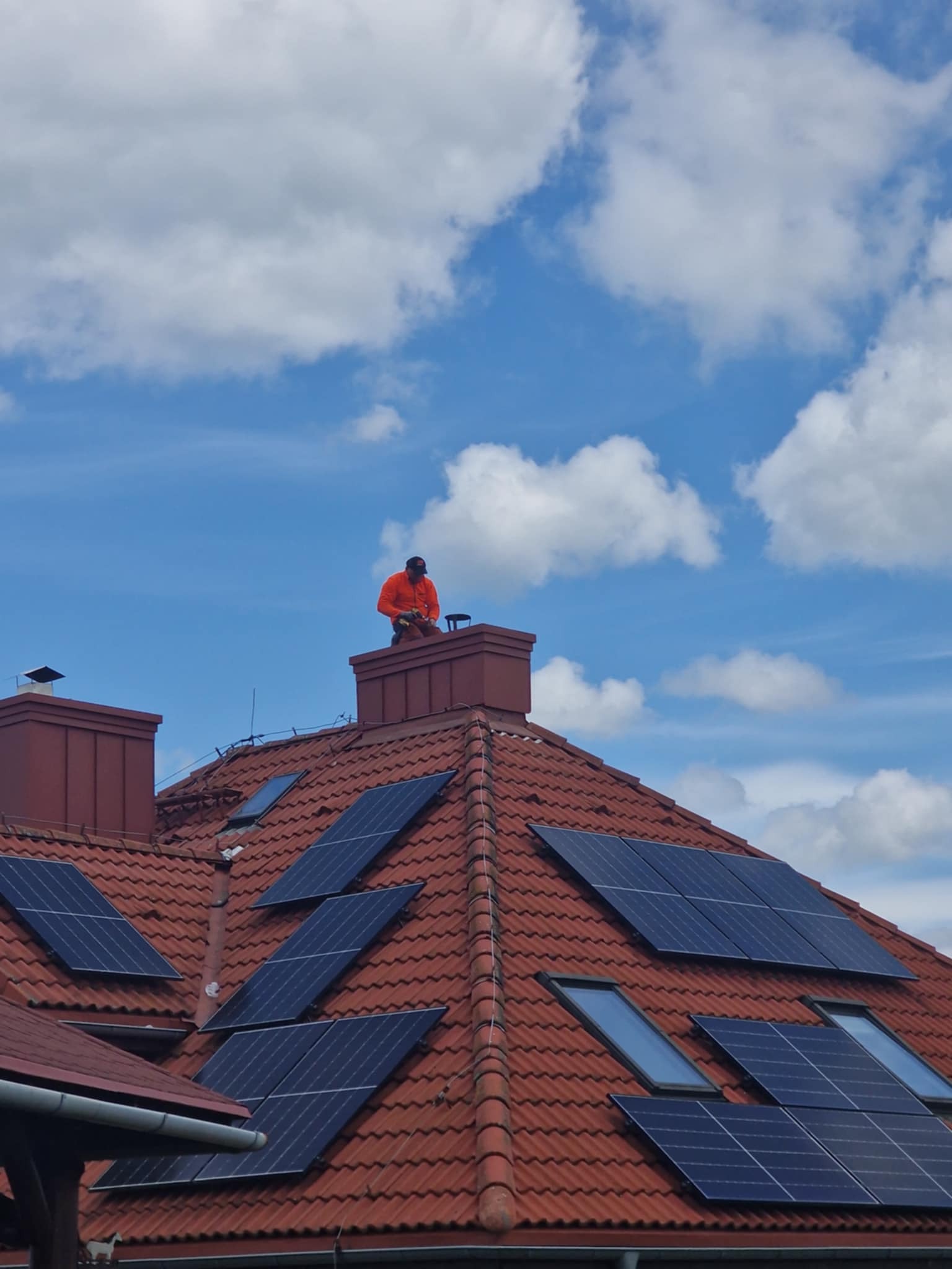 Czerwony dach z panelami słonecznymi i oknami połaciowymi w Milówce. Na kominie dekarz w pomarańczowej kurtce. Błękitne niebo z białymi chmurami w tle.