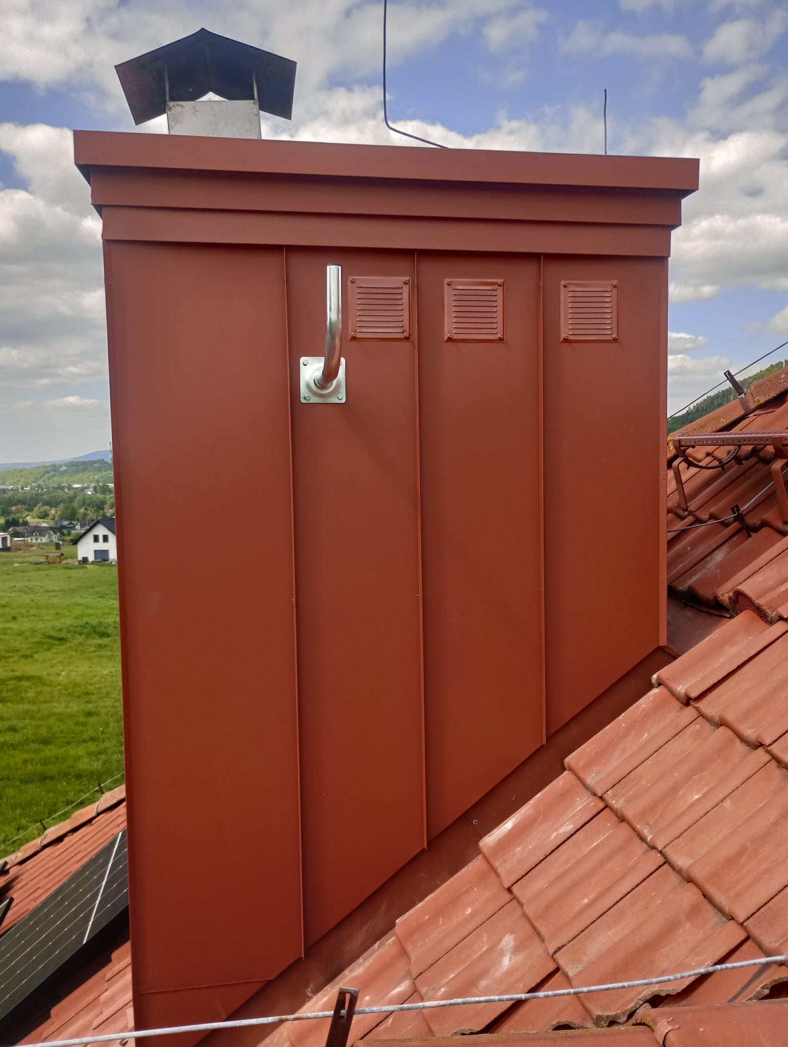 Czerwono-brązowa obróbka blacharska komina z trzema kratkami wentylacyjnymi, na tle dachu z czerwonej dachówki i krajobrazu z zielonymi polami.