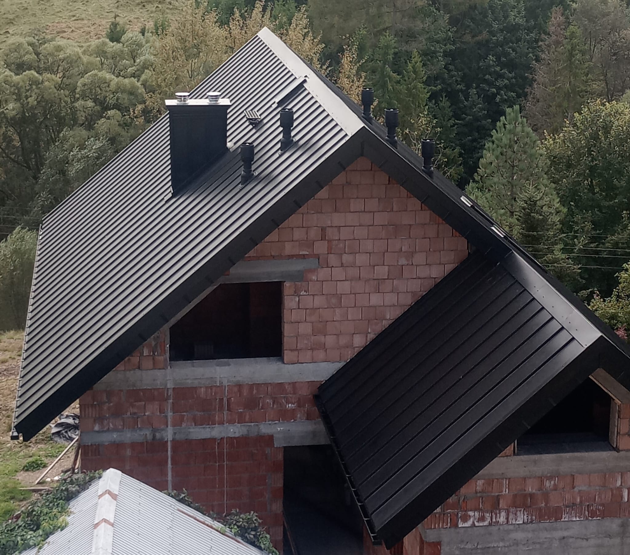 Dom w trakcie budowy z czarnym dachem z blachy na tle zieleni. Widoczne kominy i ceglana konstrukcja budynku. Dach pokryty blachą trapezową.