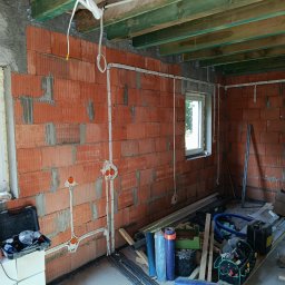 Marcin - Wnętrze w trakcie budowy z widocznymi czerwonymi ścianami z pustaków, ułożonymi białymi peszlami instalacji elektrycznej i pomarańczowymi puszkami. Zielone belki stropowe i narzędzia na podłodze.