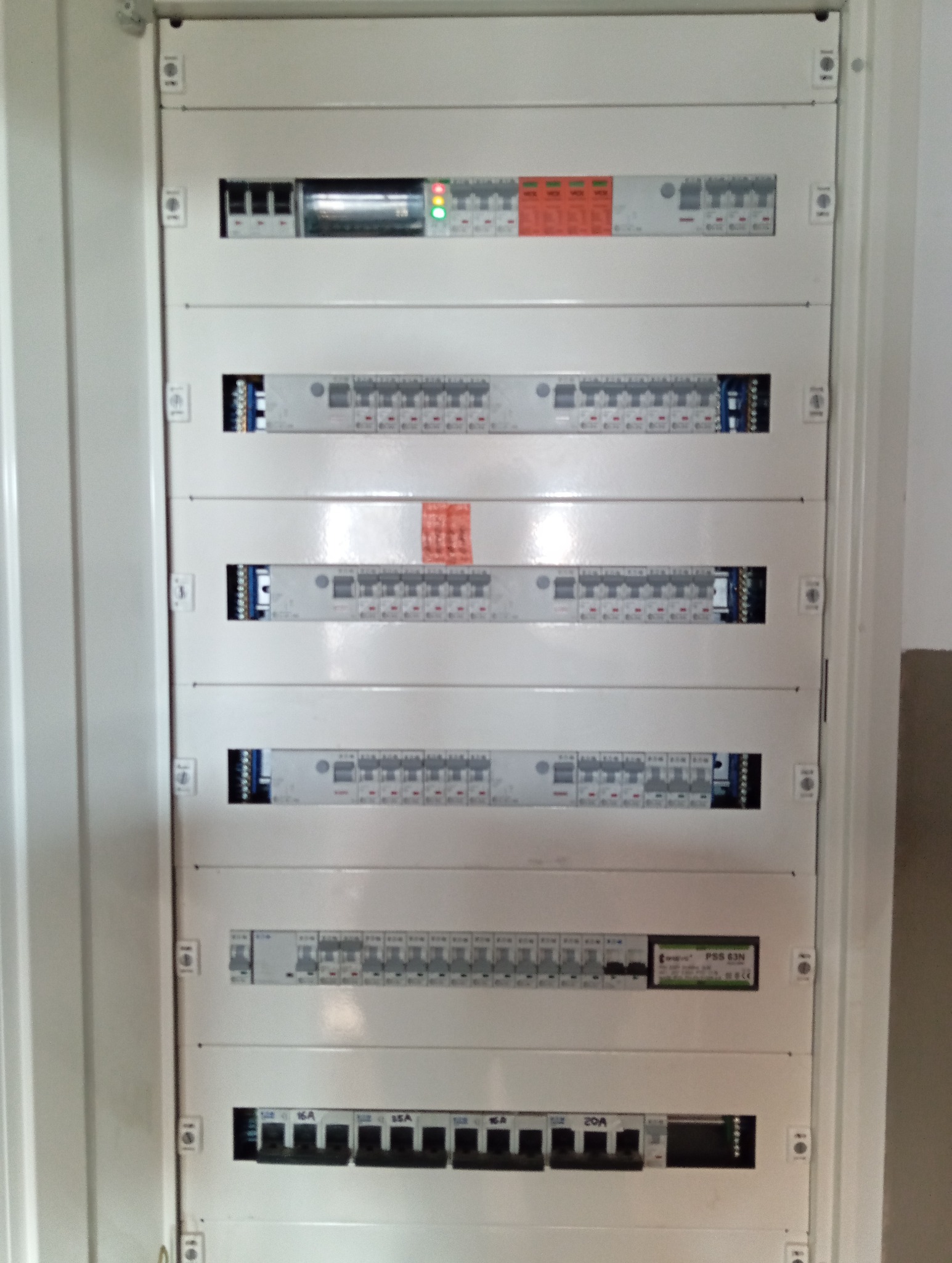 Rozdzielnica elektryczna z bezpiecznikami i wyłącznikami, widok z bliska. Biała obudowa, liczne moduły zabezpieczeń, oznaczenia amperażu.