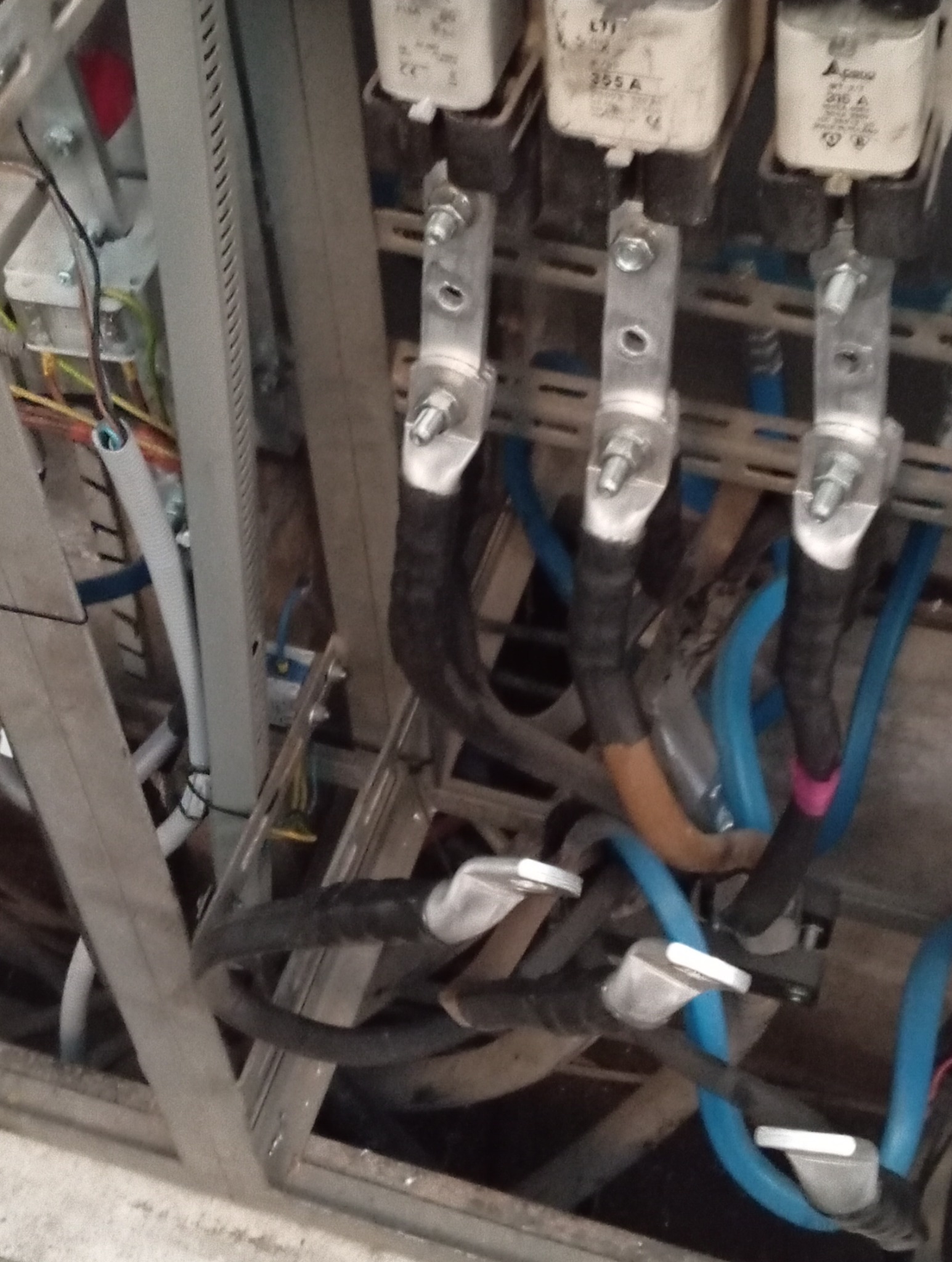 Szczegółowe ujęcie rozdzielnicy elektrycznej z widocznymi bezpiecznikami, przewodami w izolacji i metalową konstrukcją wsporczą. Elementy instalacji w kolorach czarnym i niebieskim.