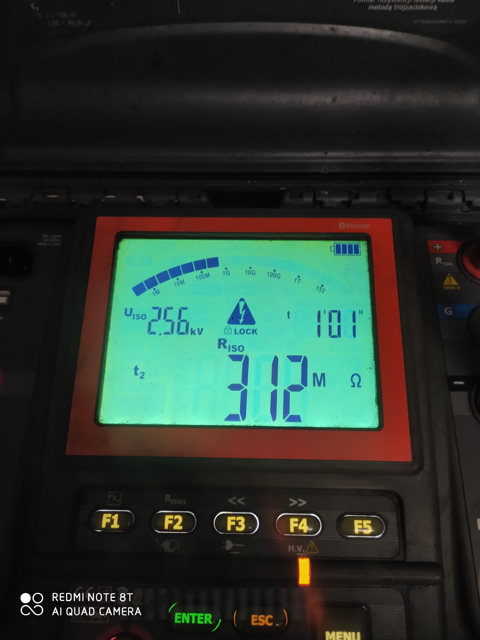Miernik rezystancji izolacji z podświetlonym ekranem LCD, wskazujący wartości pomiarowe napięcia 2.56kV i rezystancji 312MΩ. Widoczne przyciski funkcyjne i oznaczenia.