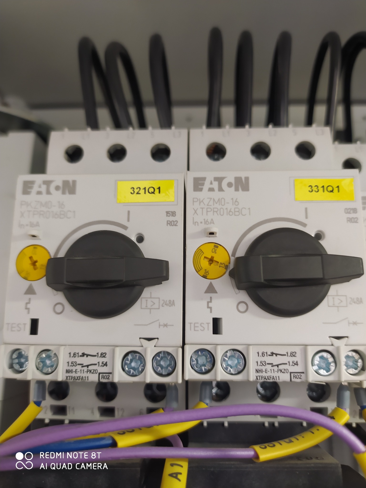 Dwa identyczne, białe wyłączniki Eaton PKZM0-16 z czarnymi pokrętłami, opisane żółtymi etykietami 321Q1 i 331Q1, z podłączonymi przewodami w kolorach żółtym i fioletowym.