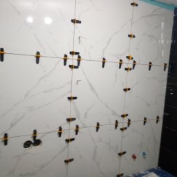 Gabbud - Ściana w trakcie układania płytek imitujących marmur, z czarnymi krzyżykami dystansowymi i otworami na instalacje, sąsiadująca z fragmentem ściany wyłożonej ciemnymi płytkami