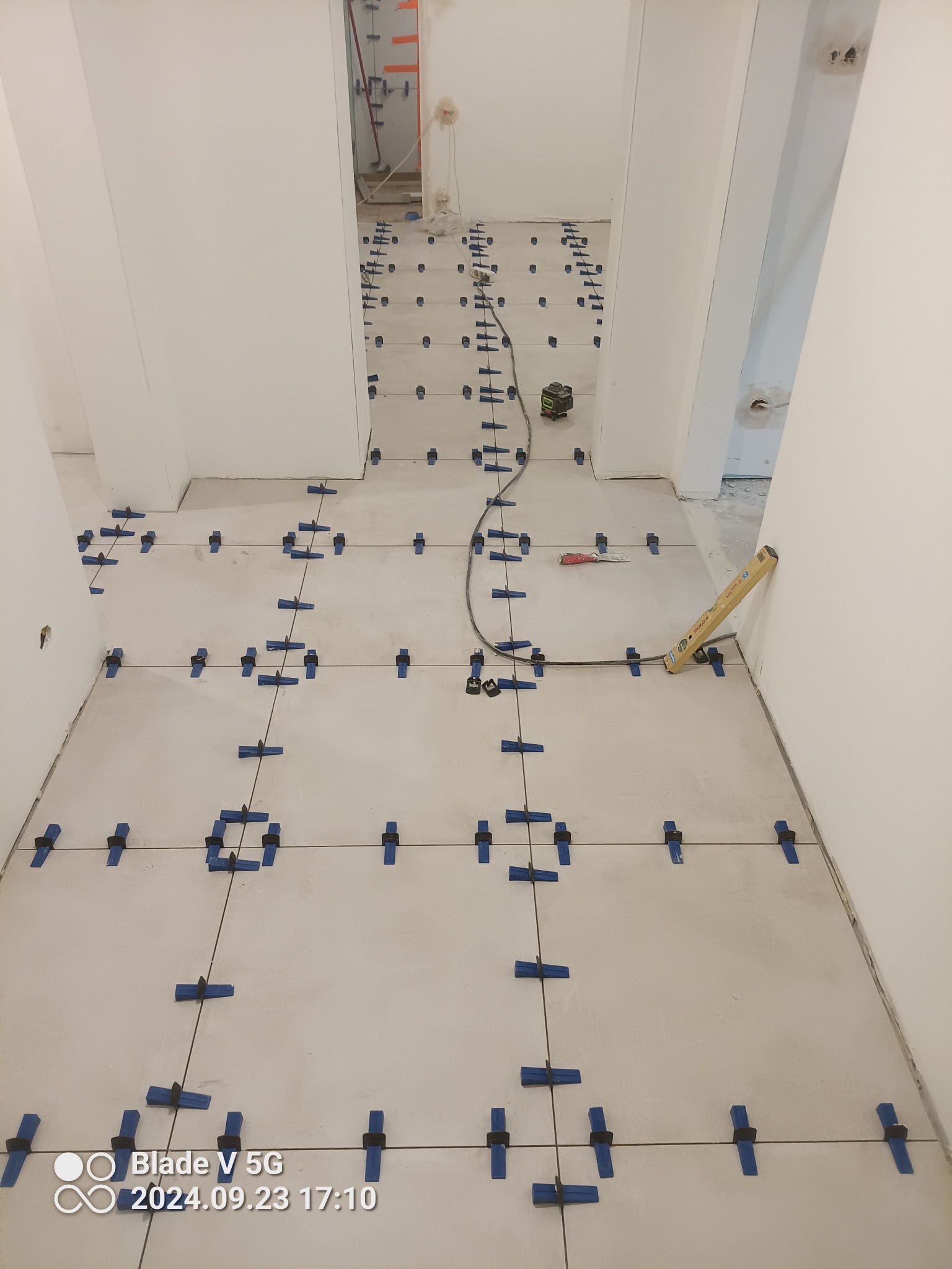 Układanie jasnych płytek podłogowych w korytarzu z użyciem niebieskich klipsów poziomujących, widoczny laserowy niwelator i poziomica przy ścianie.