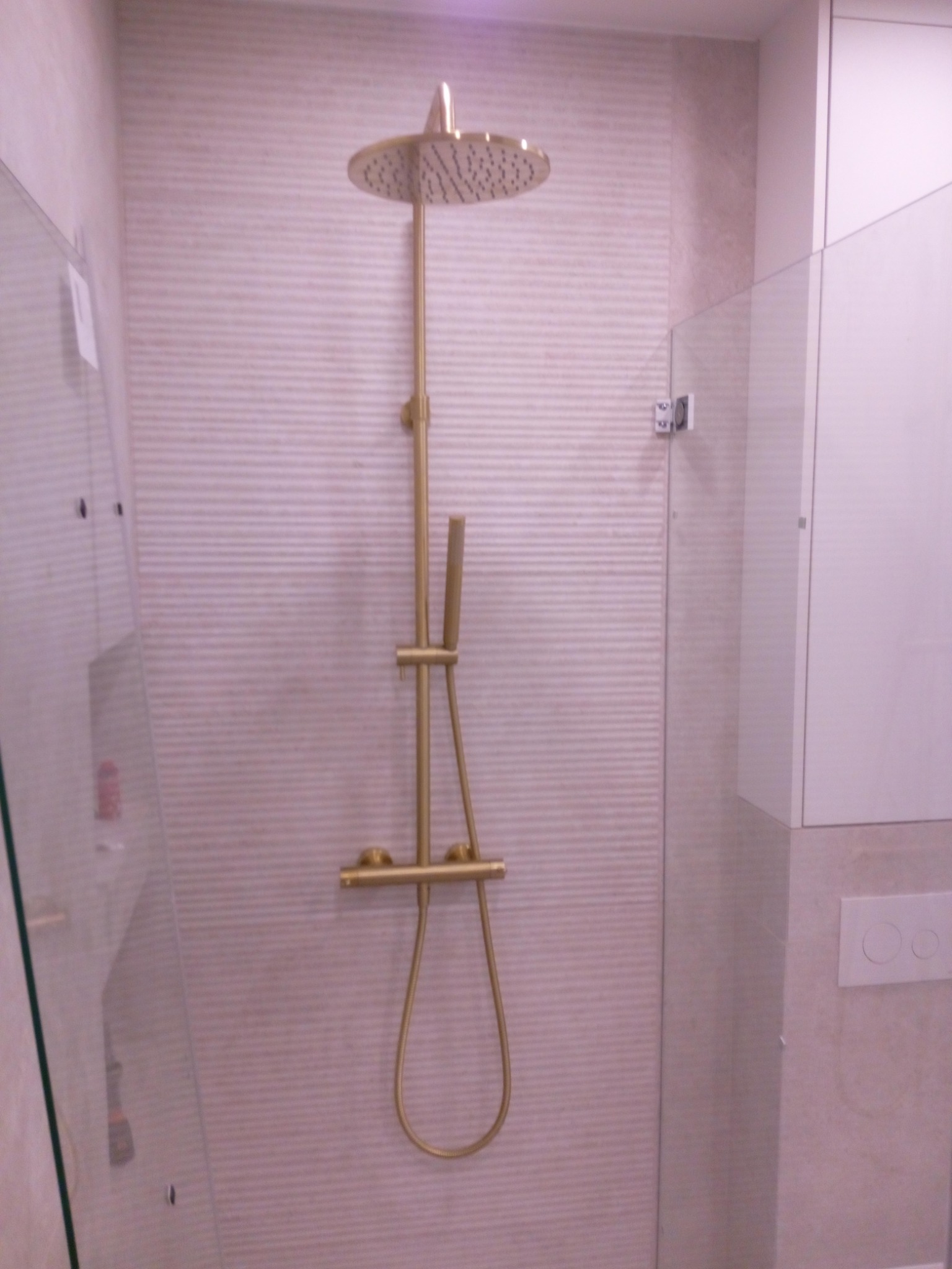 Nowoczesna kabina prysznicowa z deszczownicą w kolorze złotym, jasne płytki z delikatnym wzorem poziomych pasków, minimalistyczny design.