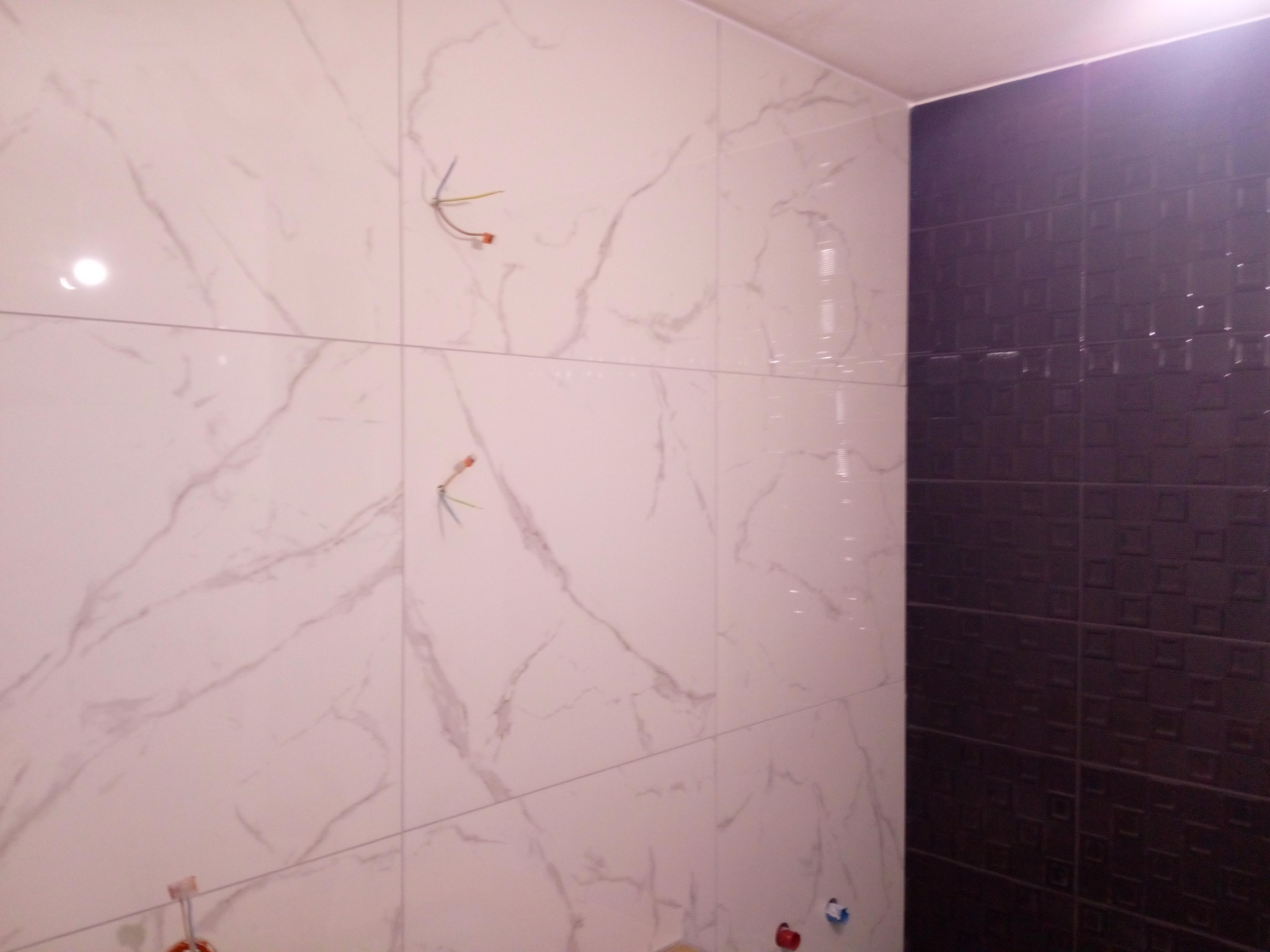 Ściana łazienki wyłożona białymi płytkami imitującymi marmur, z widocznymi wystającymi przewodami elektrycznymi i rurami wodnymi przed montażem armatury, sąsiadująca z ciemną ścianą z płytek...