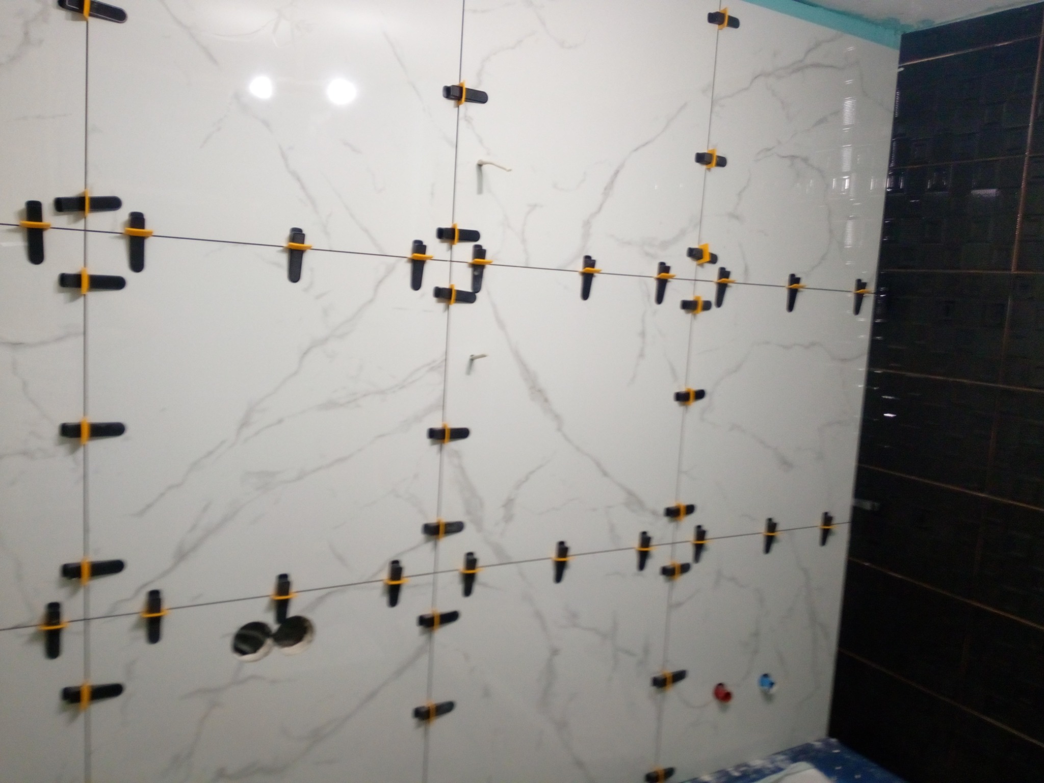 Ściana w trakcie układania płytek imitujących marmur, z czarnymi krzyżykami dystansowymi i otworami na instalacje, sąsiadująca z fragmentem ściany wyłożonej ciemnymi płytkami