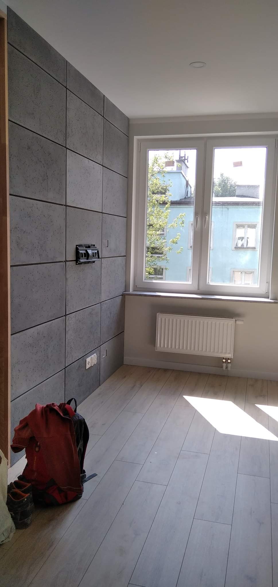 Pokój po remoncie: szare panele ścienne imitujące beton, częściowo zamontowane gniazdka elektryczne, jasne panele podłogowe, okno z widokiem na budynek, czerwony plecak i buty robocze w rogu.