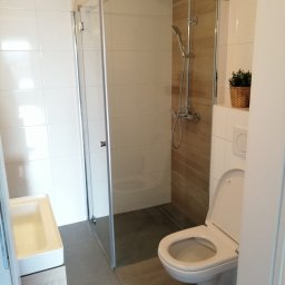 Paweł Kozłowski - Nowoczesna łazienka z kabiną prysznicową ze szklanymi drzwiami, wiszącą toaletą z otwartą klapą i umywalką z szafką, wykończona białymi i drewnopodobnymi płytkami, z doniczką na półce.