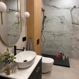 Nowoczesna łazienka z marmurowymi płytkami, owalną umywalką na szafce z ryflowanymi frontami i złotymi uchwytami, dekoracją świąteczną i kabiną prysznicową typu walk-in z czarną armaturą.