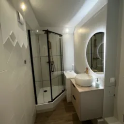 Nowoczesna łazienka z kabiną prysznicową typu walk-in z czarnymi profilami, okrągłym lustrem z podświetleniem LED i umywalką nablatową.