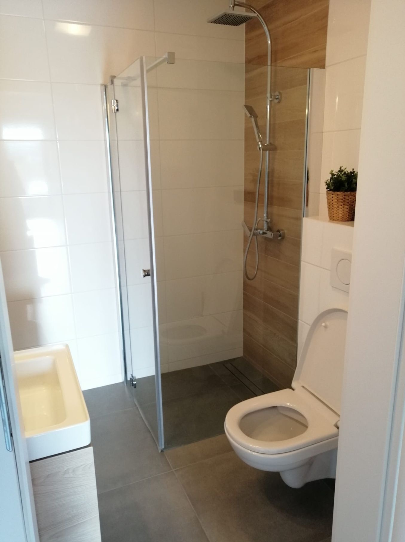 Nowoczesna łazienka z kabiną prysznicową ze szklanymi drzwiami, wiszącą toaletą z otwartą klapą i umywalką z szafką, wykończona białymi i drewnopodobnymi płytkami, z doniczką na półce.