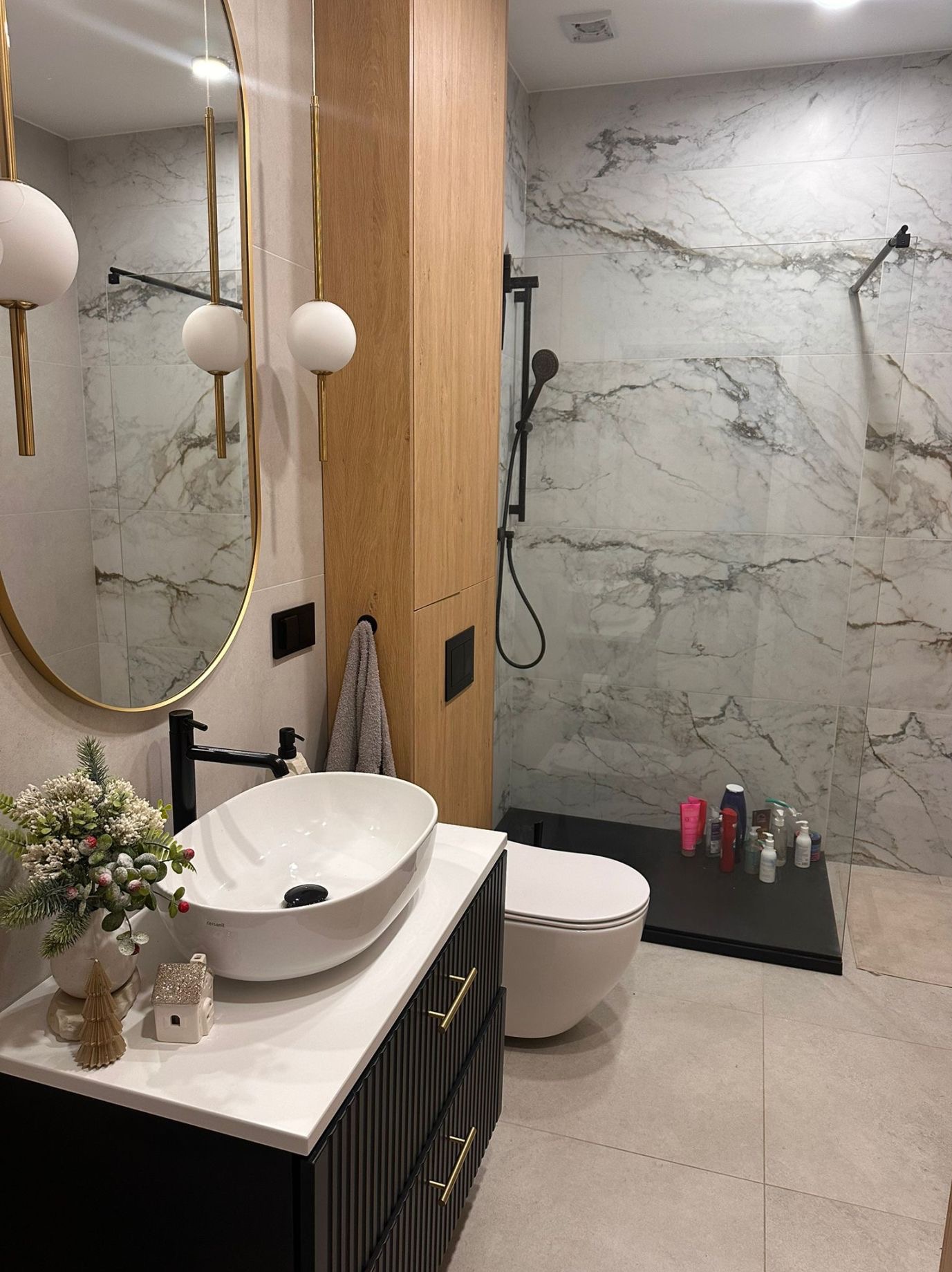 Nowoczesna łazienka z marmurowymi płytkami, owalną umywalką na szafce z ryflowanymi frontami i złotymi uchwytami, dekoracją świąteczną i kabiną prysznicową typu walk-in z czarną armaturą.