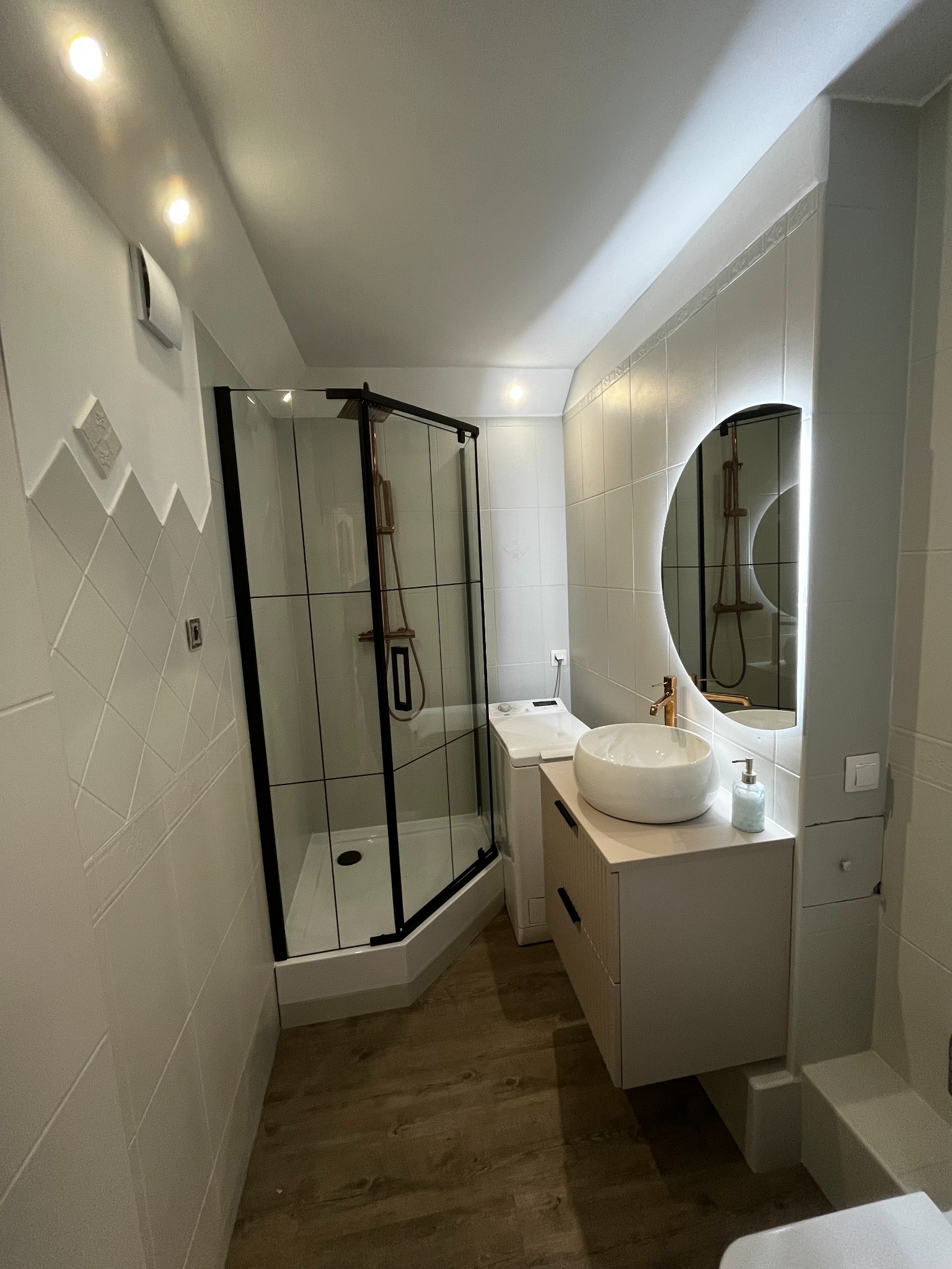 Nowoczesna łazienka z kabiną prysznicową typu walk-in z czarnymi profilami, okrągłym lustrem z podświetleniem LED i umywalką nablatową.