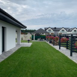 GARDEMO Projektowanie i zakładanie terenów zielonych Marcin Olszak - Starannie utrzymany trawnik przylegający do nowoczesnego domu z szarą kostką brukową, widoczne czarne ogrodzenie z wiszącymi donicami z różowymi kwiatami i altana w tle, w tle zabudowa szeregowa.