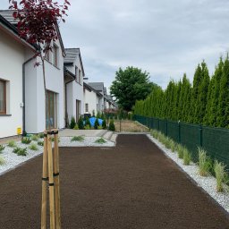 GARDEMO Projektowanie i zakładanie terenów zielonych Marcin Olszak - Przygotowany teren pod trawnik z widocznym systemem nawadniania, obrzeżami z białego kamienia i nasadzeniami wzdłuż ogrodzenia oraz drzewkiem z purpurowymi liśćmi zabezpieczonym drewnianymi palikami.