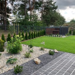 GARDEMO Projektowanie i zakładanie terenów zielonych Marcin Olszak - Uporządkowany ogród z równo przystrzyżonym trawnikiem, ozdobnymi kamieniami i roślinami, widok z perspektywy kostki brukowej, w tle rząd tuj i drewniana altana.
