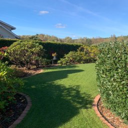 GARDEMO Projektowanie i zakładanie terenów zielonych Marcin Olszak - Zadbany trawnik z równo przystrzyżoną trawą, obrzeża z czerwonej cegły, krzewy i żywopłot w tle, mały stolik z doniczką w zacienionym miejscu ogrodu.