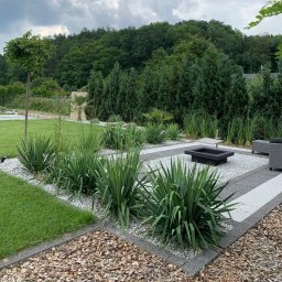 GARDEMO Projektowanie i zakładanie terenów zielonych Marcin Olszak - Nowoczesny ogród z geometrycznym układem ścieżek z białego i szarego żwiru, obrzeżami z kory i nasadzeniami wysokich traw oraz juk, w tle drzewa iglaste i liściaste, czarne palenisko i szare meble...