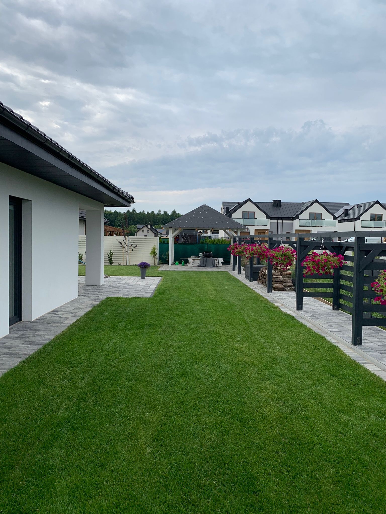 Starannie utrzymany trawnik przylegający do nowoczesnego domu z szarą kostką brukową, widoczne czarne ogrodzenie z wiszącymi donicami z różowymi kwiatami i altana w tle, w tle zabudowa szeregowa.