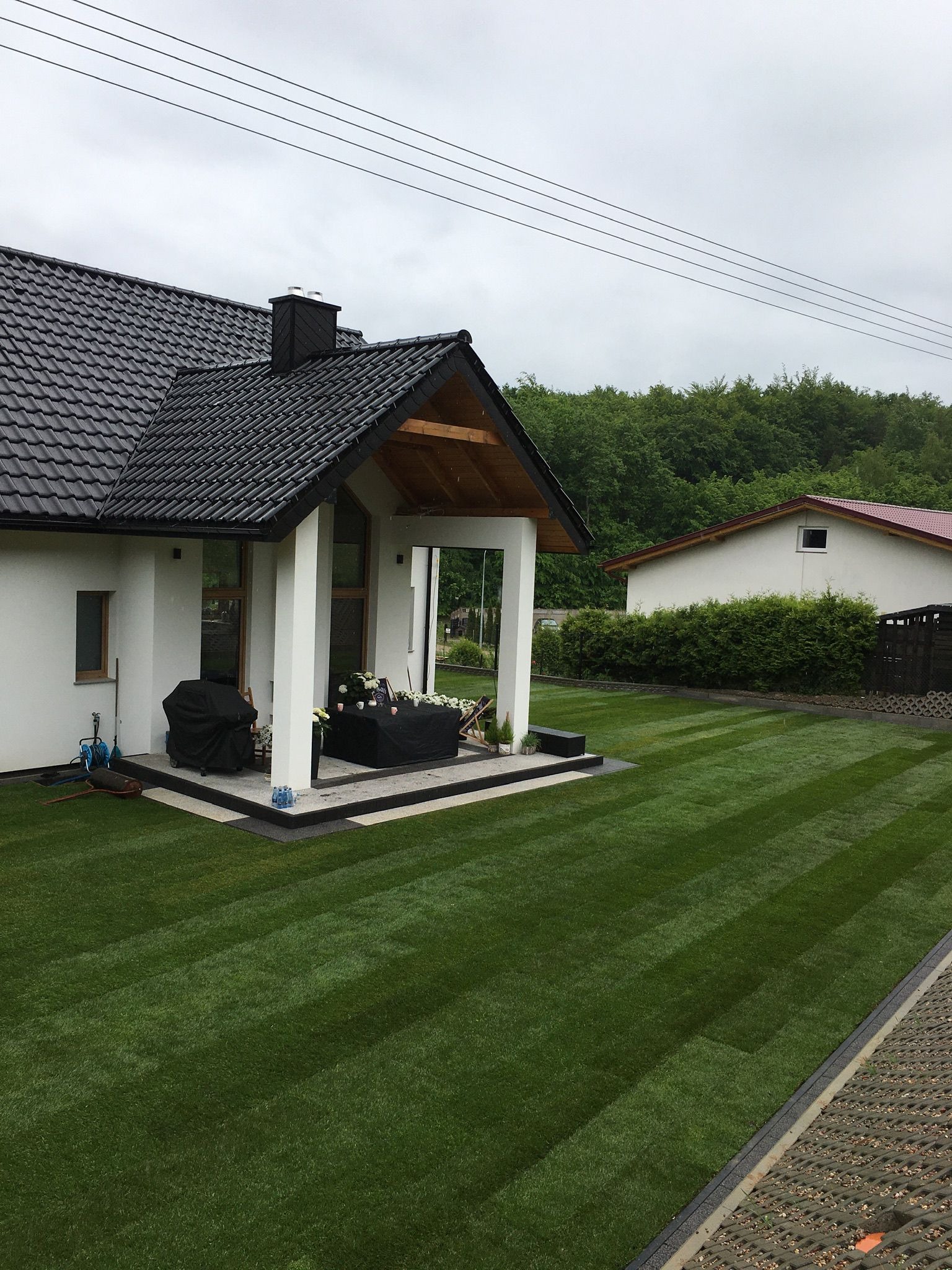 Perfekcyjnie przystrzyżony trawnik z wyraźnymi pasami, przylegający do nowoczesnego domu z zadaszonym tarasem, gdzie ustawiono dekoracje i sprzęt przykryty pokrowcem. W tle widoczny drugi dom...