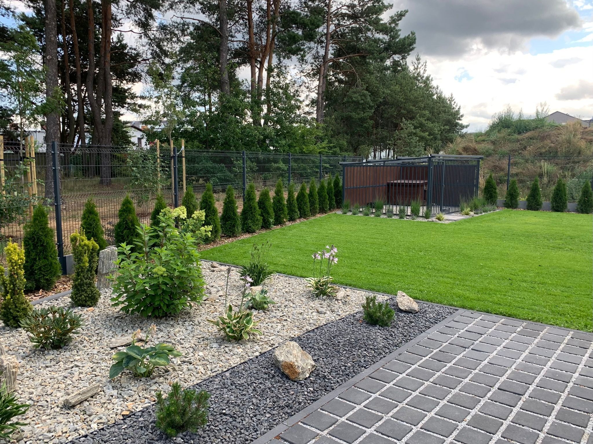 Uporządkowany ogród z równo przystrzyżonym trawnikiem, ozdobnymi kamieniami i roślinami, widok z perspektywy kostki brukowej, w tle rząd tuj i drewniana altana.