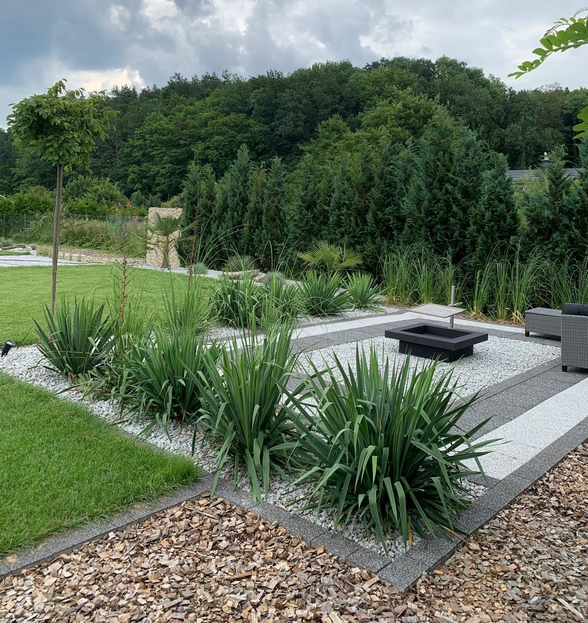 Nowoczesny ogród z geometrycznym układem ścieżek z białego i szarego żwiru, obrzeżami z kory i nasadzeniami wysokich traw oraz juk, w tle drzewa iglaste i liściaste, czarne palenisko i szare meble...