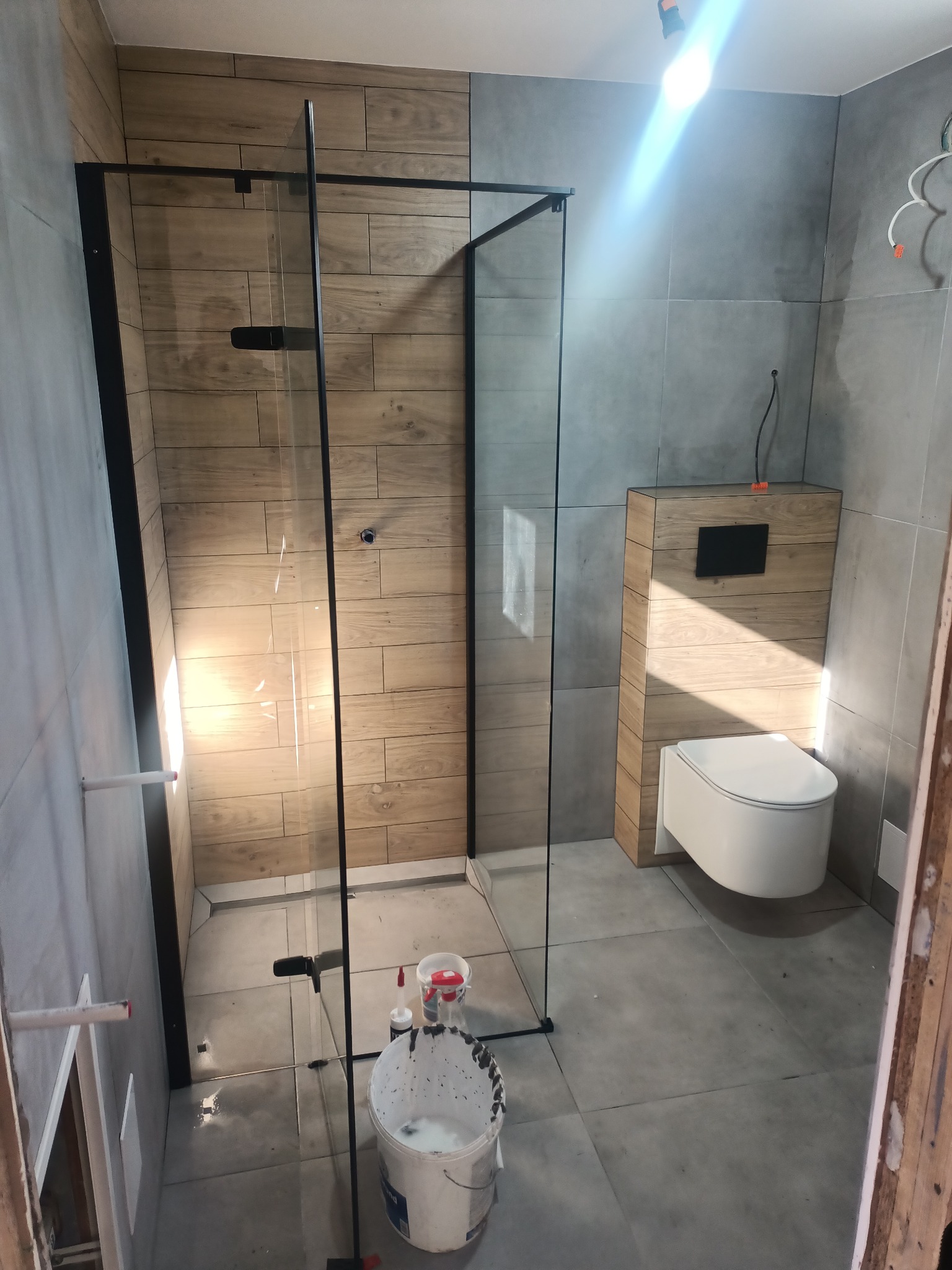 Niedokończona łazienka z kabiną prysznicową z czarnymi profilami, podwieszaną toaletą w obudowie z drewnopodobnych paneli, szare płytki na ścianach i podłodze, widoczne narzędzia i wiadro z farbą.