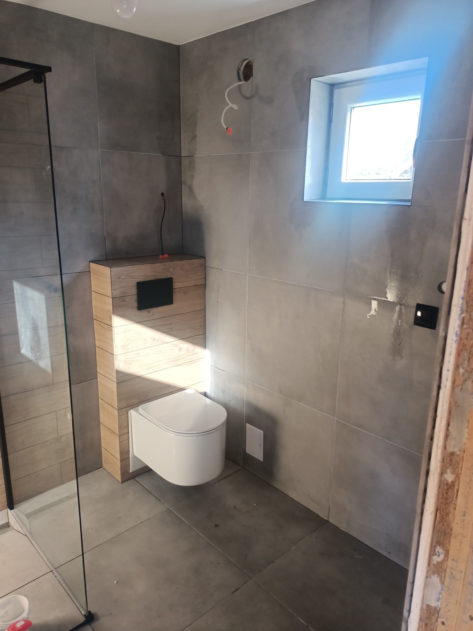 Szara łazienka w trakcie prac wykończeniowych z podwieszaną toaletą z drewnianą obudową, widoczne ślady po instalacjach elektrycznych, zamontowane okno i fragment kabiny prysznicowej.